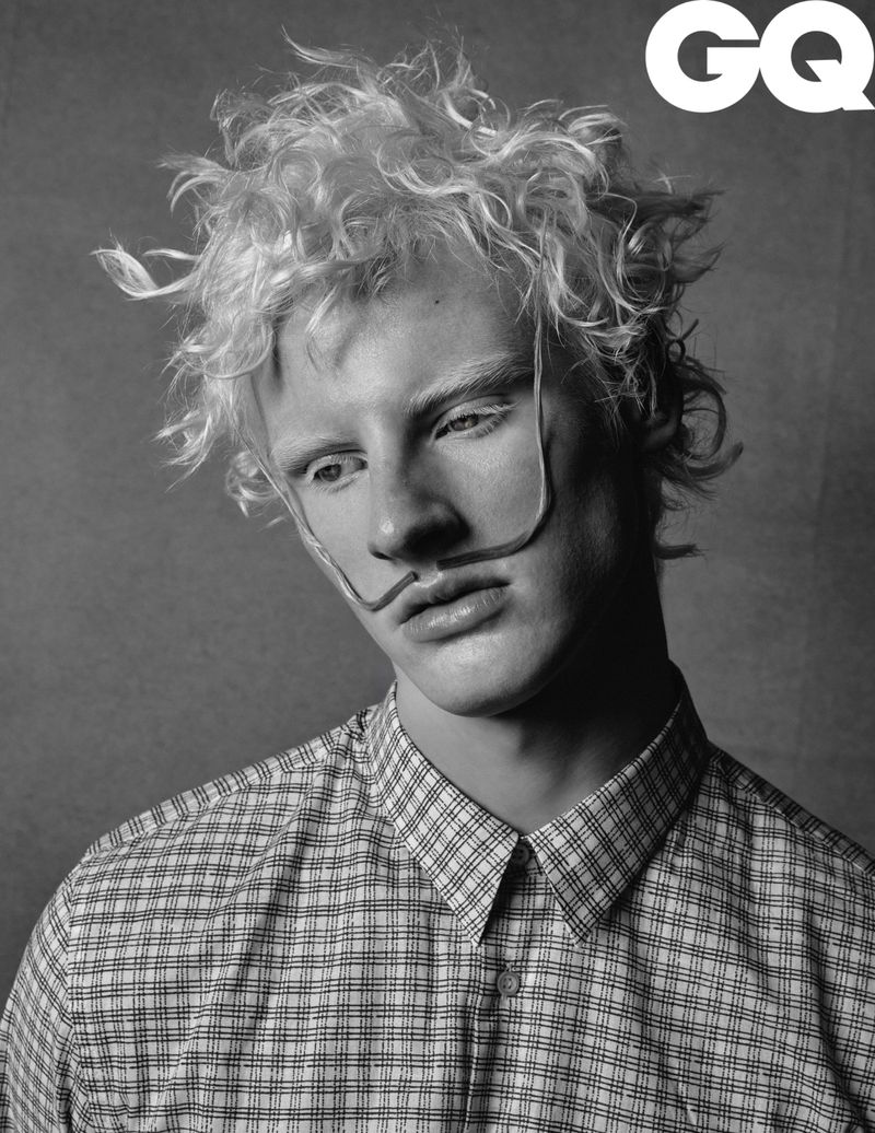 Monochrome portrait of a young adult man with textured hair and facial decorations, wearing a plaid button-down shirt, GQ logo visible.