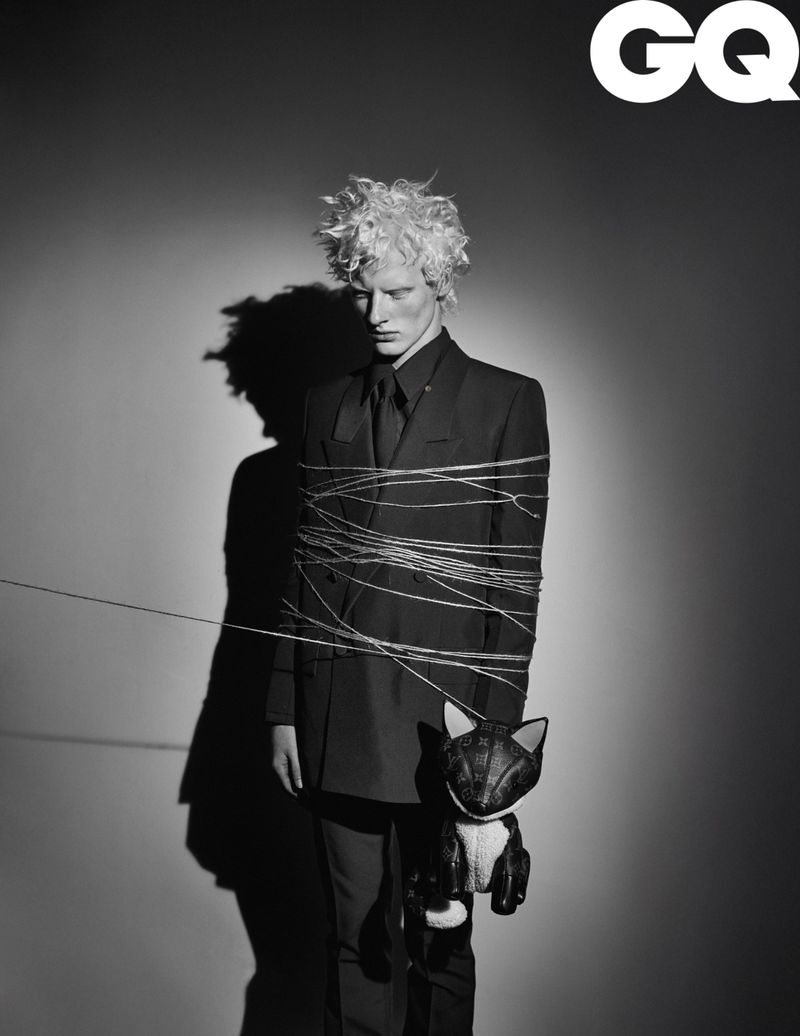 Black and white portrait of a blond male fashion model in a suit tied with string holding a cat doll in a studio for GQ Magazine.