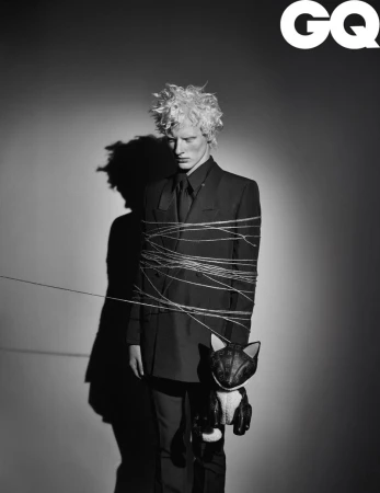 Black and white portrait of a blond male fashion model in a suit tied with string holding a cat doll in a studio for GQ Magazine.