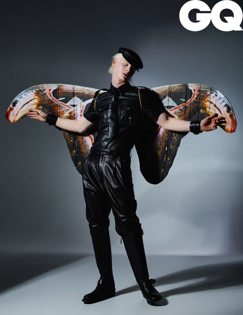 Editorial fashion shot of a fair-skinned adult male in a black leather outfit and hat, wearing decorative moth wings against a grey backdrop.