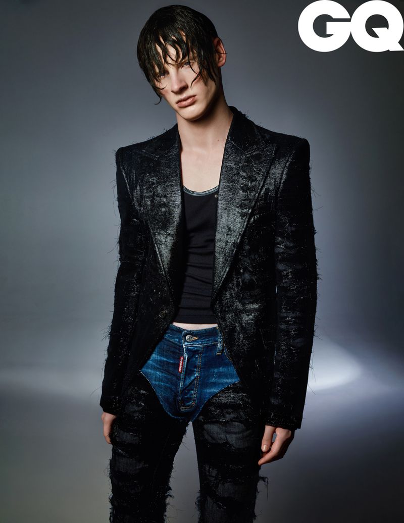 A stylish, slender male model with wet hair poses in a black distressed jacket and jeans with an inset of denim fabric, captured in a studio shot.