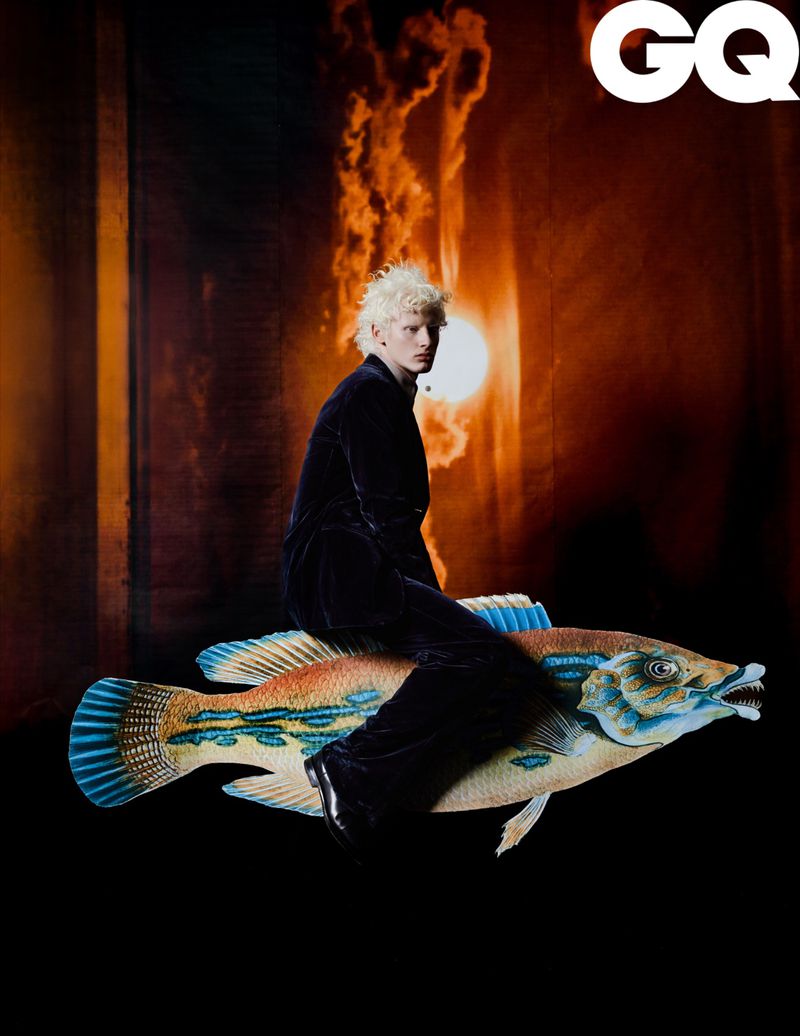 Adult male poses on an oversized fish prop in a studio with a fiery background, under GQ logo, wearing a black velvet suit.