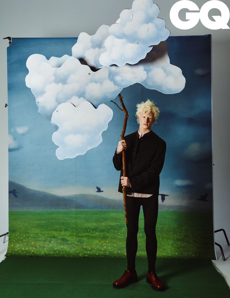 Young adult poses in front of painted backdrop holding a tree branch with cloud prop for a GQ magazine studio portrait.