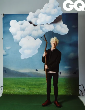 Young adult poses in front of painted backdrop holding a tree branch with cloud prop for a GQ magazine studio portrait.