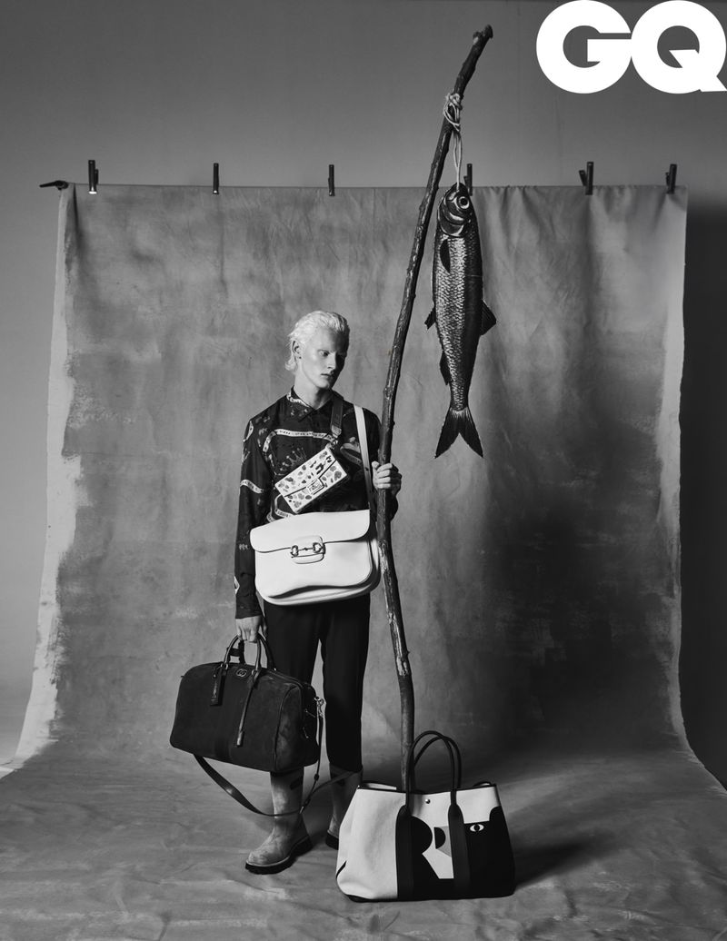 Black and white fashion shot of a non-binary person with multiple handbags, holding a branch with a hanging fish. GQ logo visible.