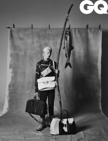 Black and white fashion shot of a non-binary person with multiple handbags, holding a branch with a hanging fish. GQ logo visible.