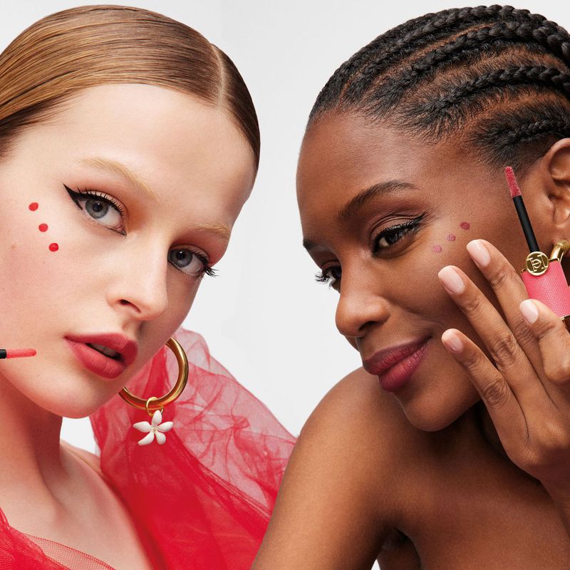 Two adult female models with red lip makeup and red dots on their faces, shot in a beauty studio with bright, even lighting.