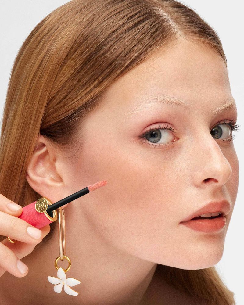 Close-up of a fair-skinned woman with red hair applying lip gloss while wearing flower earrings against a white backdrop.