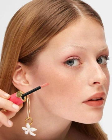 Close-up of a fair-skinned woman with red hair applying lip gloss while wearing flower earrings against a white backdrop.