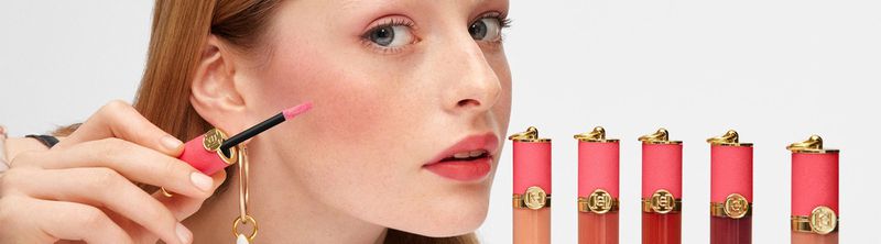 Close-up of a woman applying coral lipstick with several tubes of lipstick nearby, against a clean white backdrop in a beauty product shot.