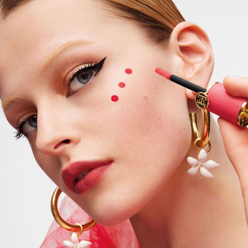 Close-up of a fair-skinned woman with red dots on her cheek. She has striking eyeliner, large gold hoop earrings, and lip product.
