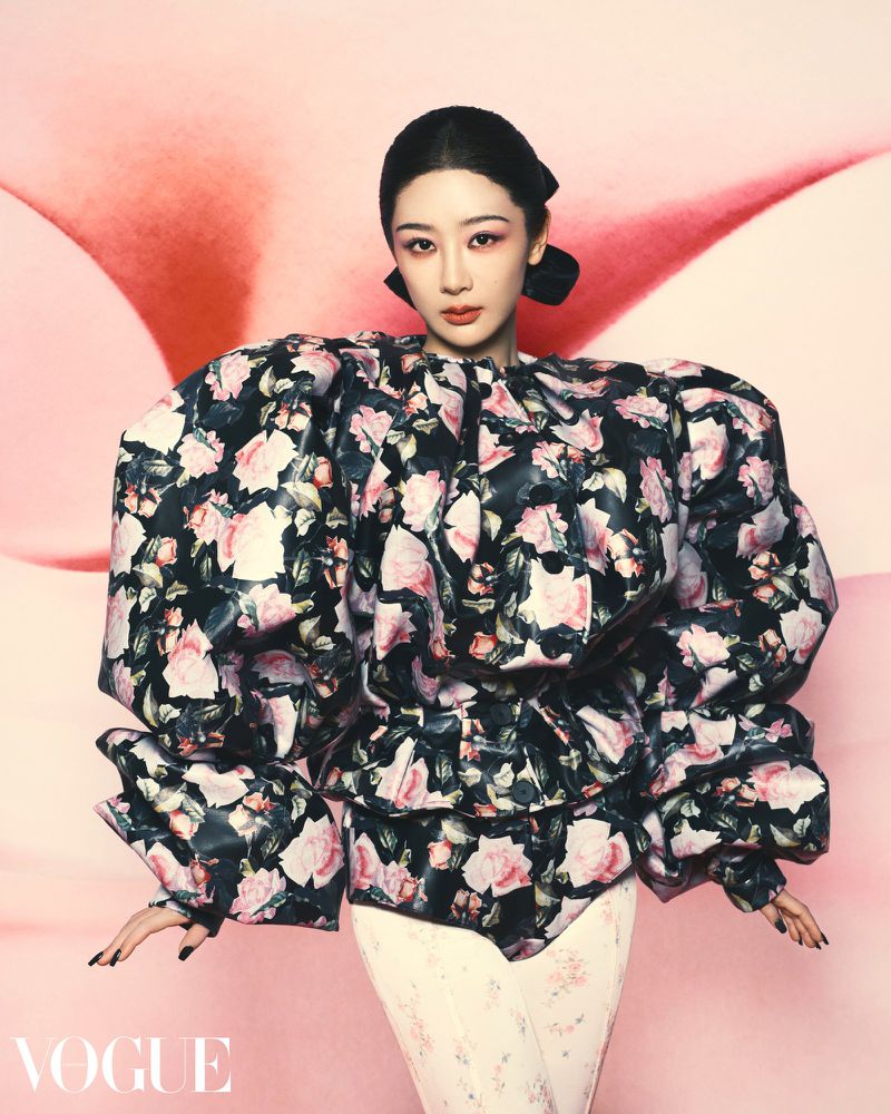 Asian woman poses in a floral top against a pink background in a fashion editorial image for Vogue magazine with a studio setup.