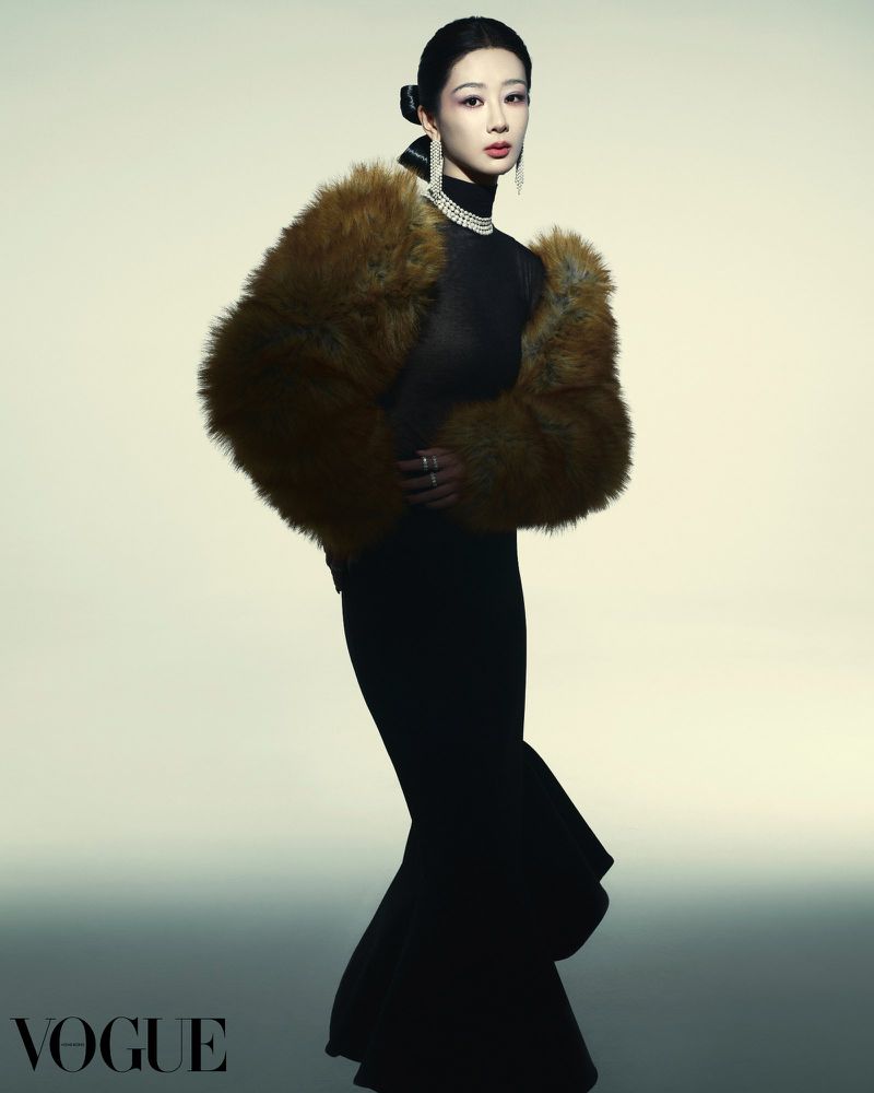 A woman in a black dress poses gracefully with a fur shawl in a fashion studio, elegantly accessorized with a pearl necklace and earrings.