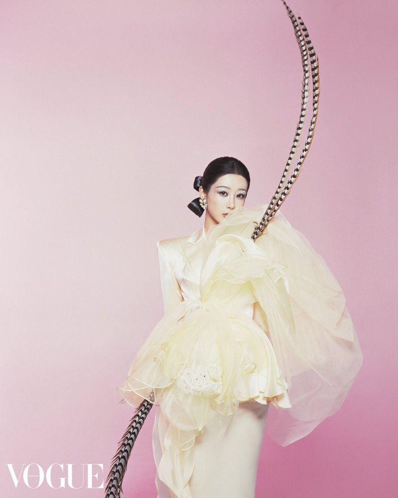 A woman poses in a light-yellow tulle dress with black-and-white striped feather accents against a pale-pink background, studio shot.