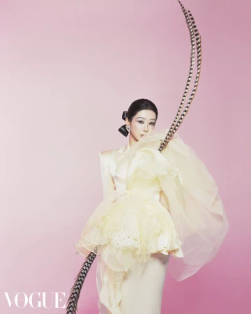 A woman poses in a light-yellow tulle dress with black-and-white striped feather accents against a pale-pink background, studio shot.