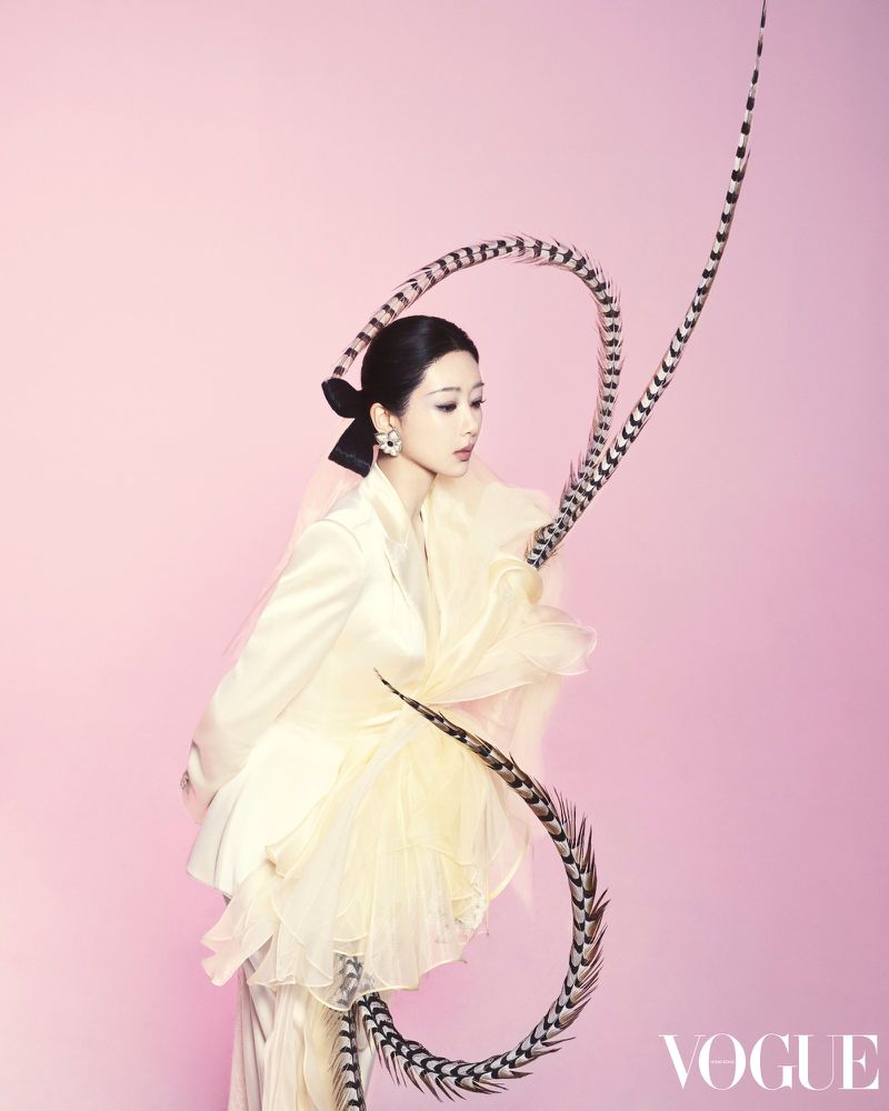 A woman poses against a pink background, wearing a white blazer, tulle accents, hair pulled back with a black bow, and feathered extensions.