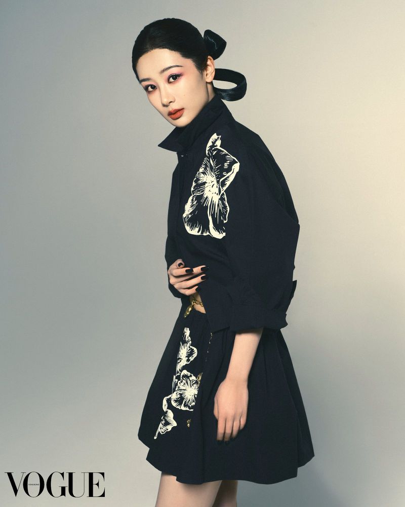 Elegant Asian woman poses in a dark, floral-patterned dress against a light gray backdrop, with the VOGUE logo in the lower left.