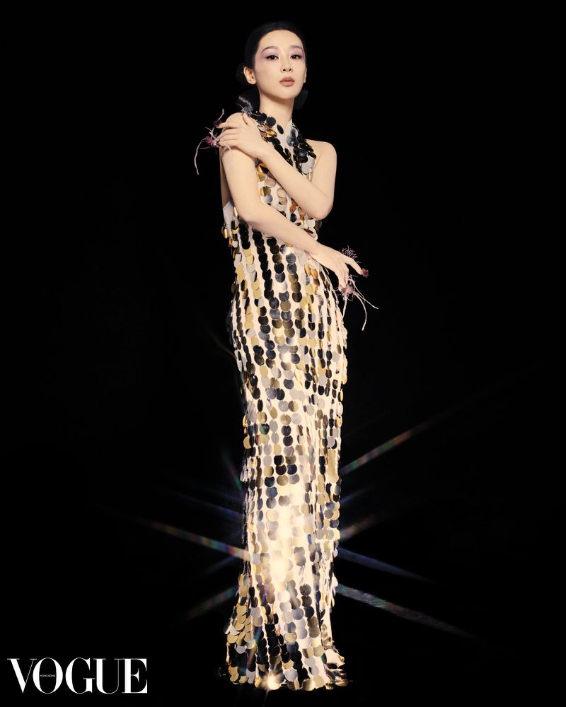 Full-length shot of a poised adult female in a sequined gown, gazing directly at the camera against a stark black backdrop, vogue logo.
