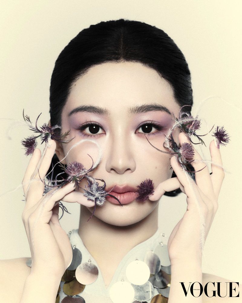 Close-up of an Asian woman with purple eye shadow and neutral lipstick posing with flowers in her hands, as seen in Vogue magazine.