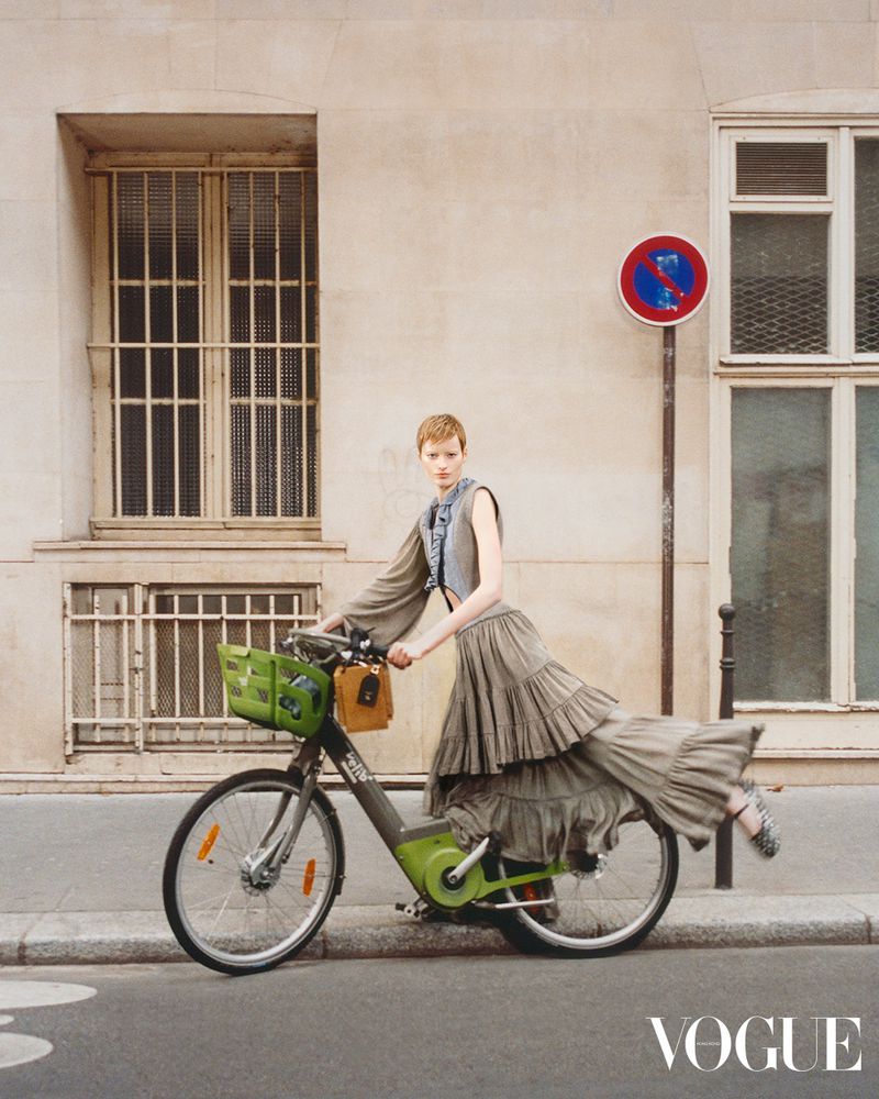 Fashion editorial shot of a young woman with short red hair in a grey dress riding a city bicycle on a city street in front of a building.