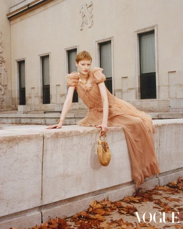 A slender adult woman sits on a stone wall in a flowing peach gown, holding a small purse, with a light stone building as backdrop.