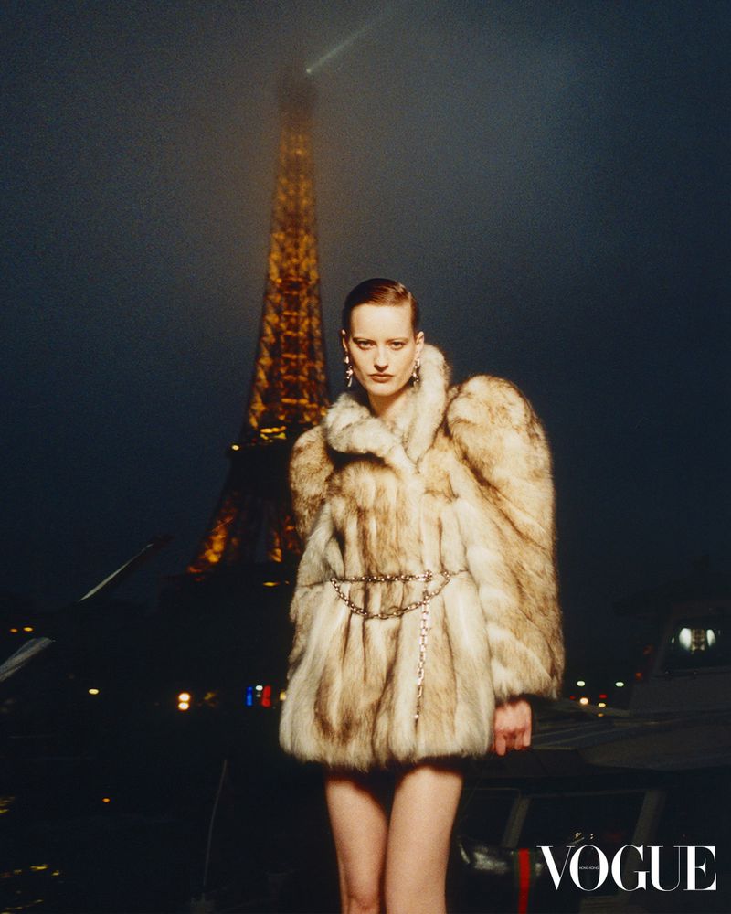 Fashion model in a fur coat poses in front of the Eiffel Tower at night for Vogue. The image showcases high-end apparel and Parisian style.