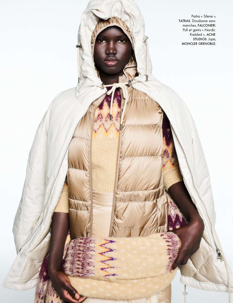 Fashion shot of a Black female model wearing a beige puffer vest, patterned top, and a matching parka with hood indoors on a white background.