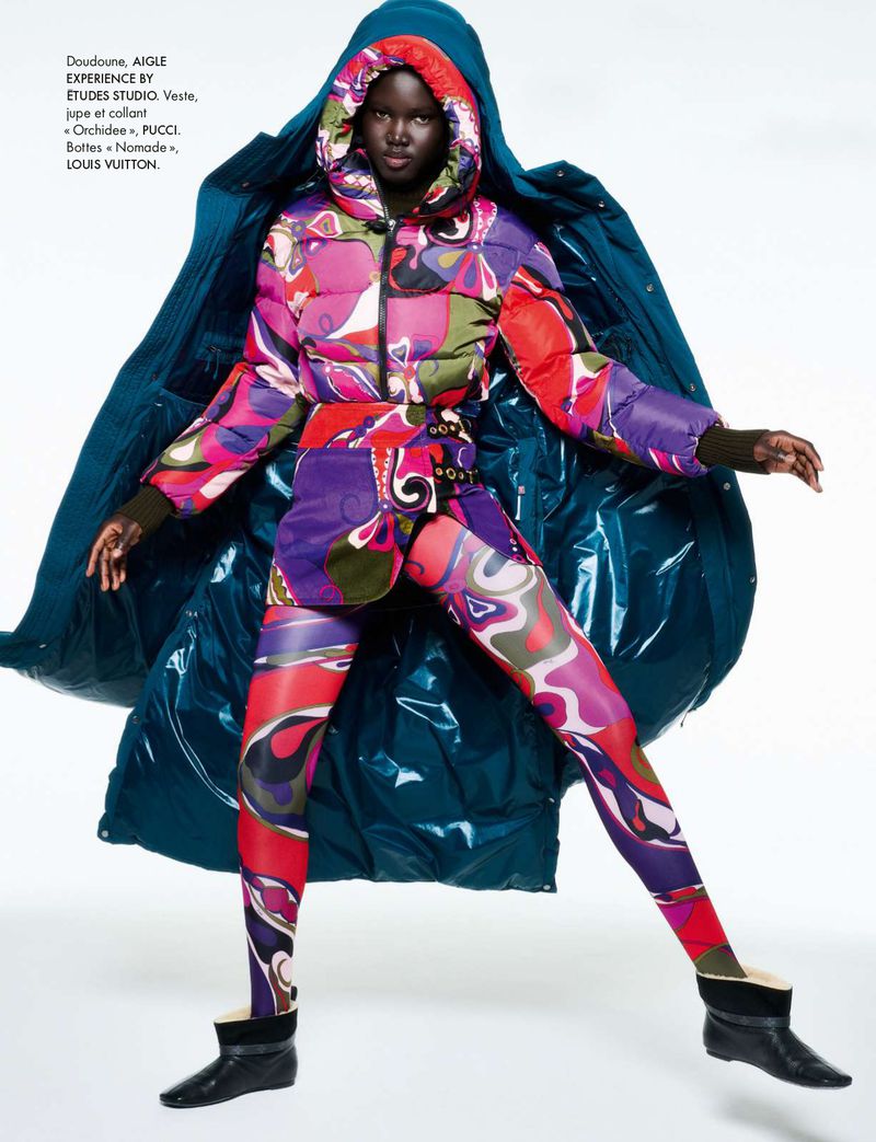 Fashion shot of an adult woman in a patterned ski suit and blue puffer coat posing against a white background in a studio.