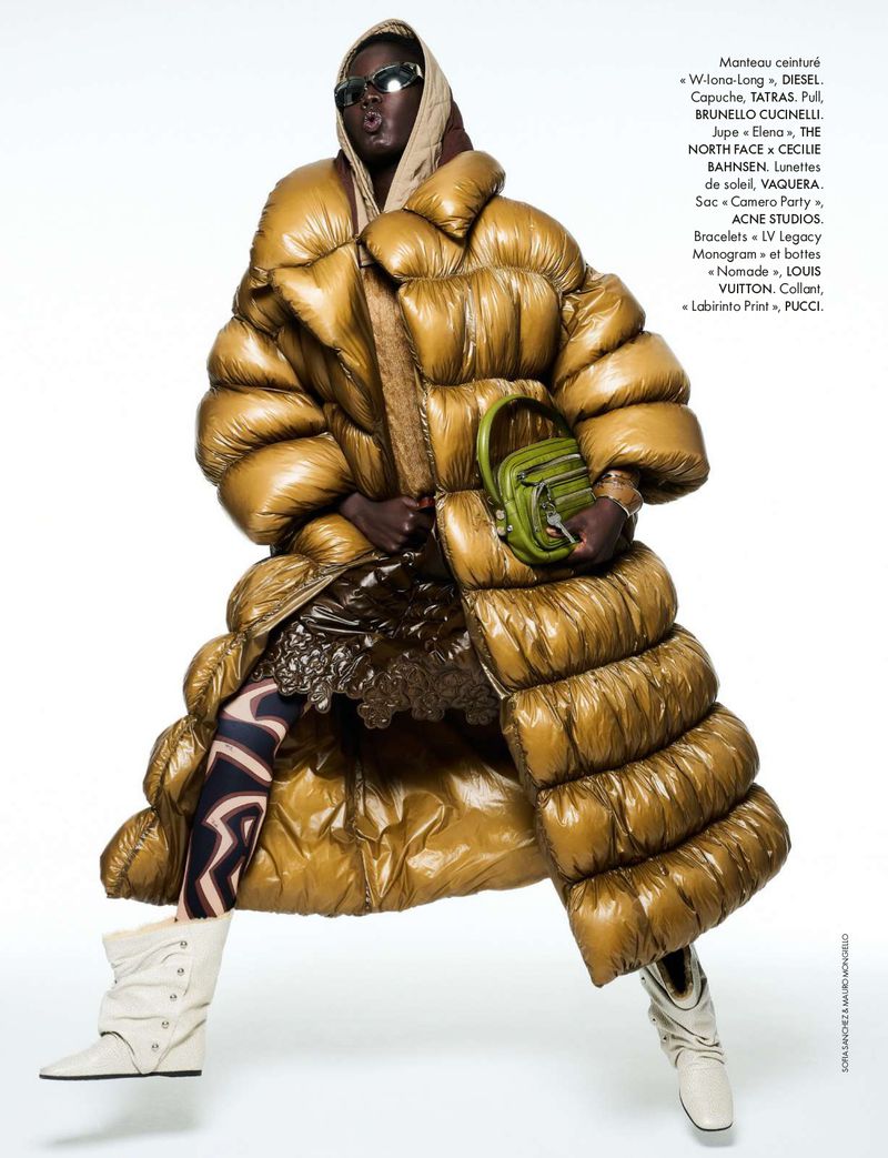 Editorial fashion shot of a woman wearing an oversized golden puffer coat with patterned leggings, boots, a headscarf, and a green purse against a white background.