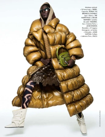 Editorial fashion shot of a woman wearing an oversized golden puffer coat with patterned leggings, boots, a headscarf, and a green purse against a white background.