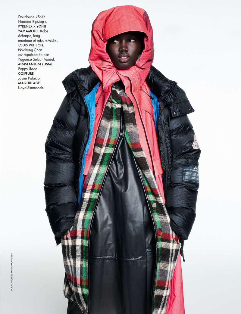 Fashion editorial shot of a Black female model wearing a black puffer coat, coral hood, and plaid scarf against a white backdrop.