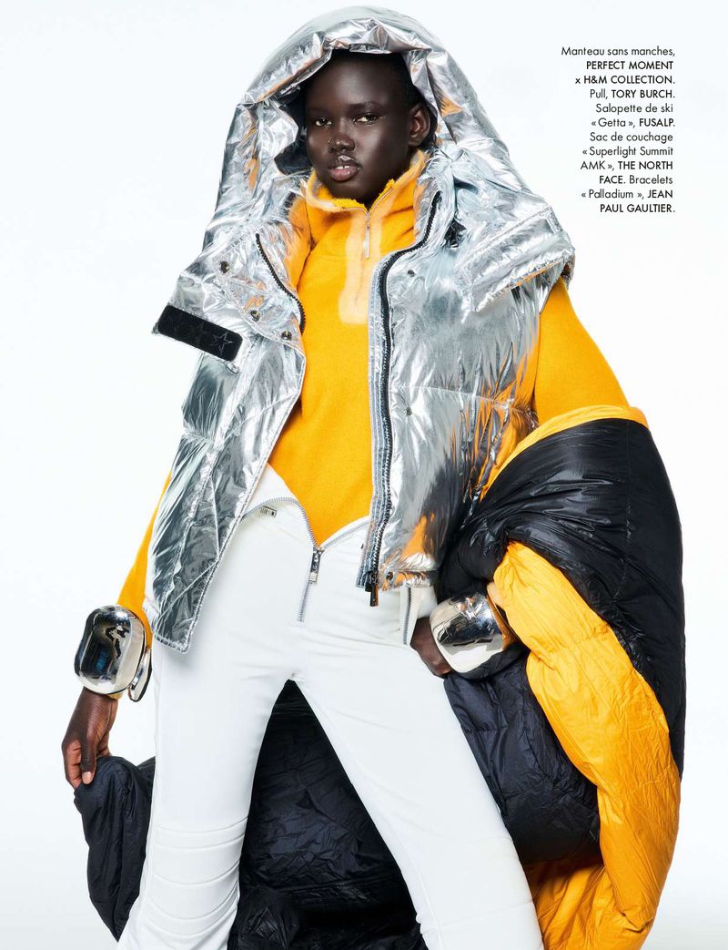 Fashion editorial shot of a Black female model in a silver hooded puffer vest, yellow top, and white pants against a white backdrop.