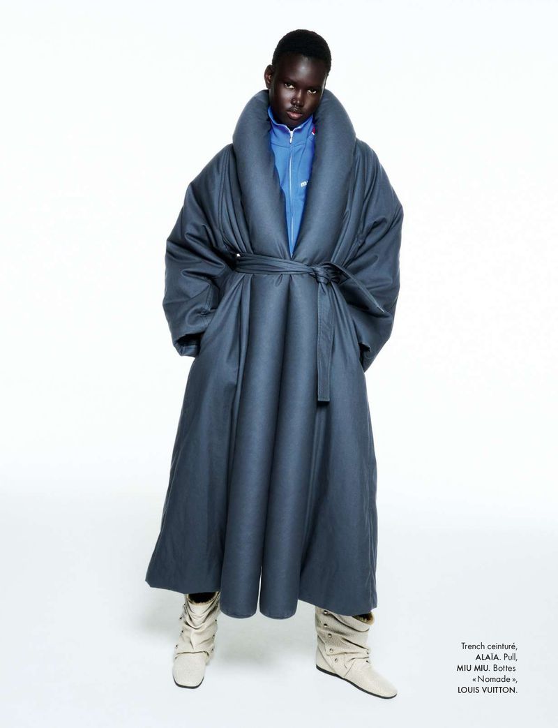 Model wearing an oversized dark blue belted coat and cream boots posed against a seamless white studio background.
