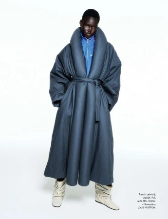 Model wearing an oversized dark blue belted coat and cream boots posed against a seamless white studio background.