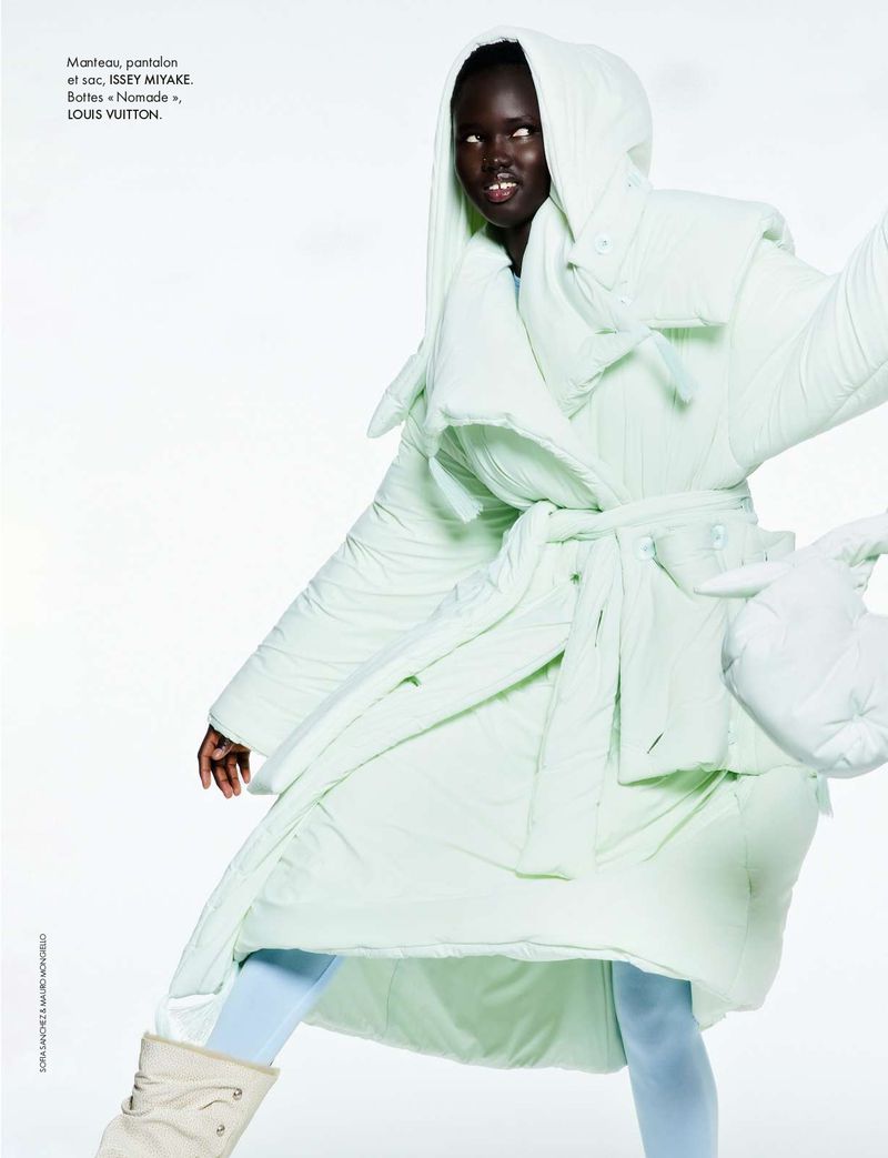 Fashion shot of a dark-skinned female model wearing a hooded, pale green puffy coat, light blue leggings, and light-colored boots on a white background.