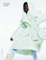 Model posing in a voluminous pale mint hooded puffer coat tied at the waist with winter boots against a white studio background.