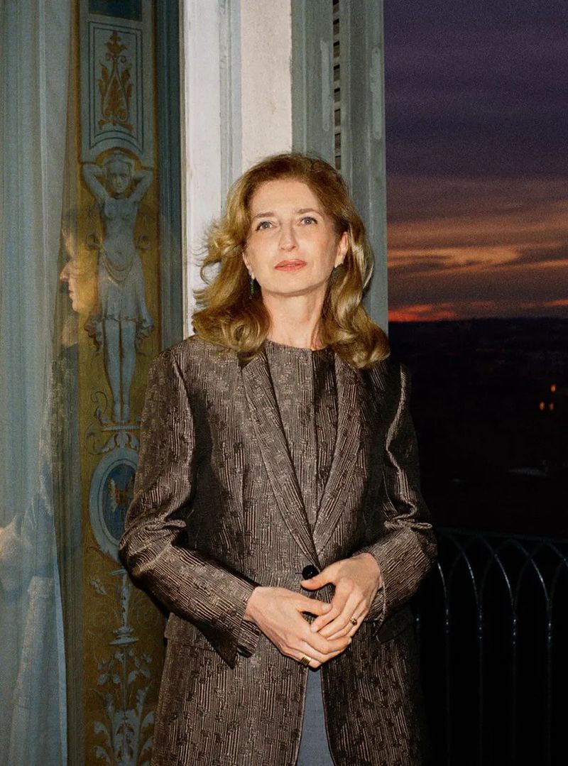 Portrait of an adult woman in a metallic blazer standing indoors near a window with a twilight view. Light hair, light skin.