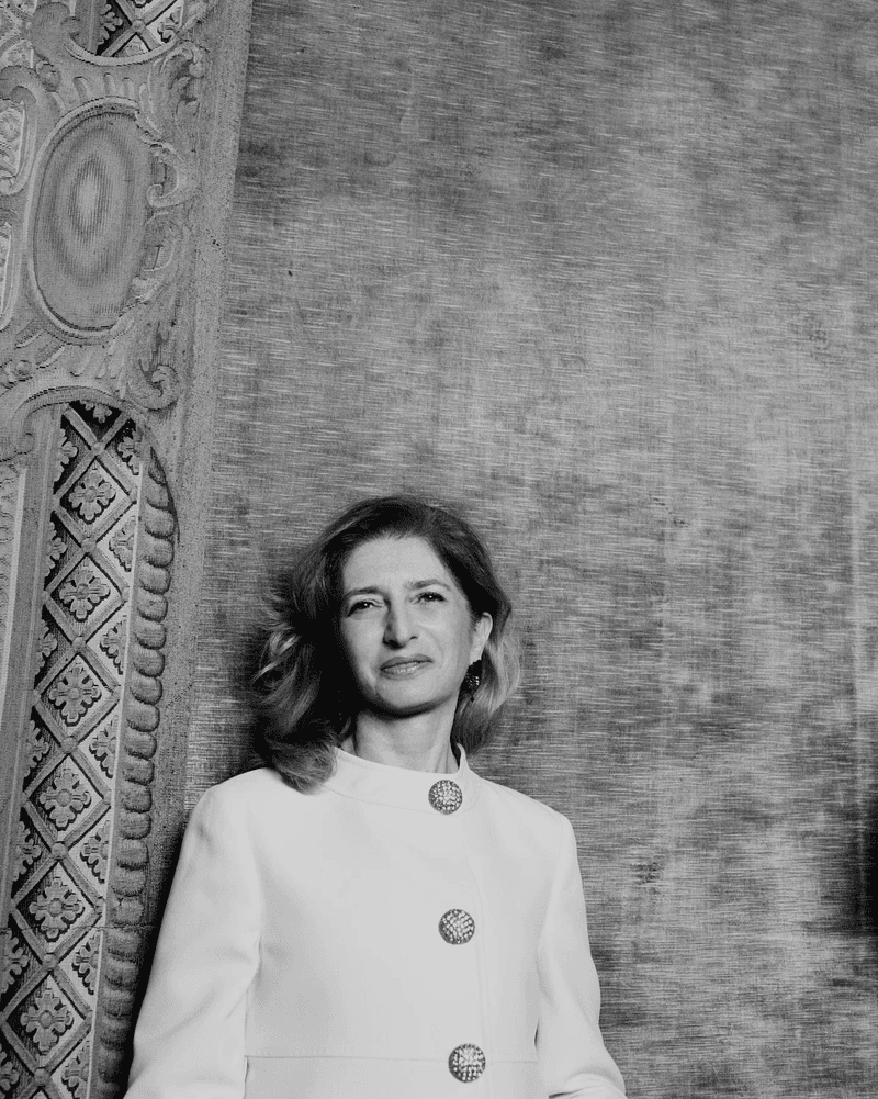 Black and white portrait of a woman in a white suit, posed against a textured wall with decorative molding on the left.