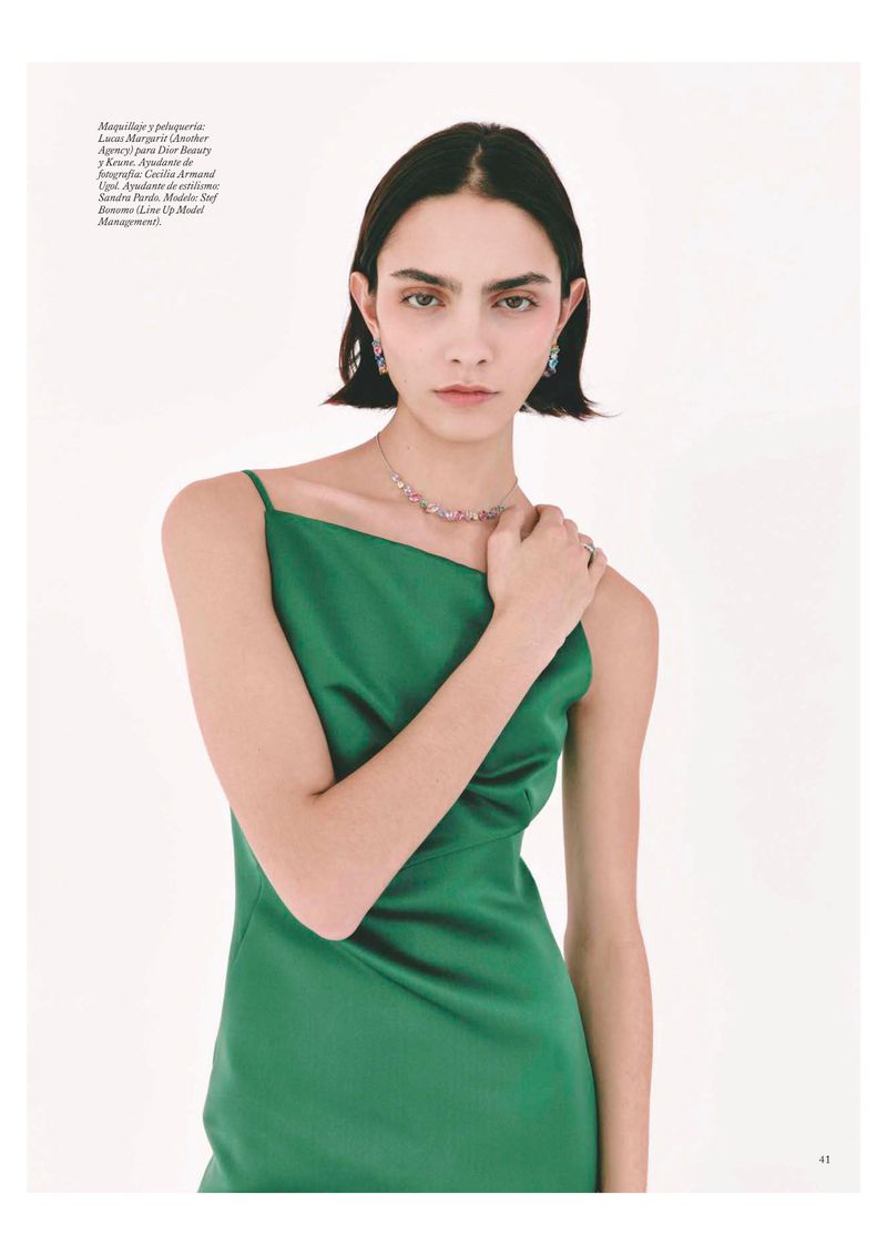 A woman with fair skin and dark hair wears a green dress and jewelry, posing in a studio with a clean, white background.