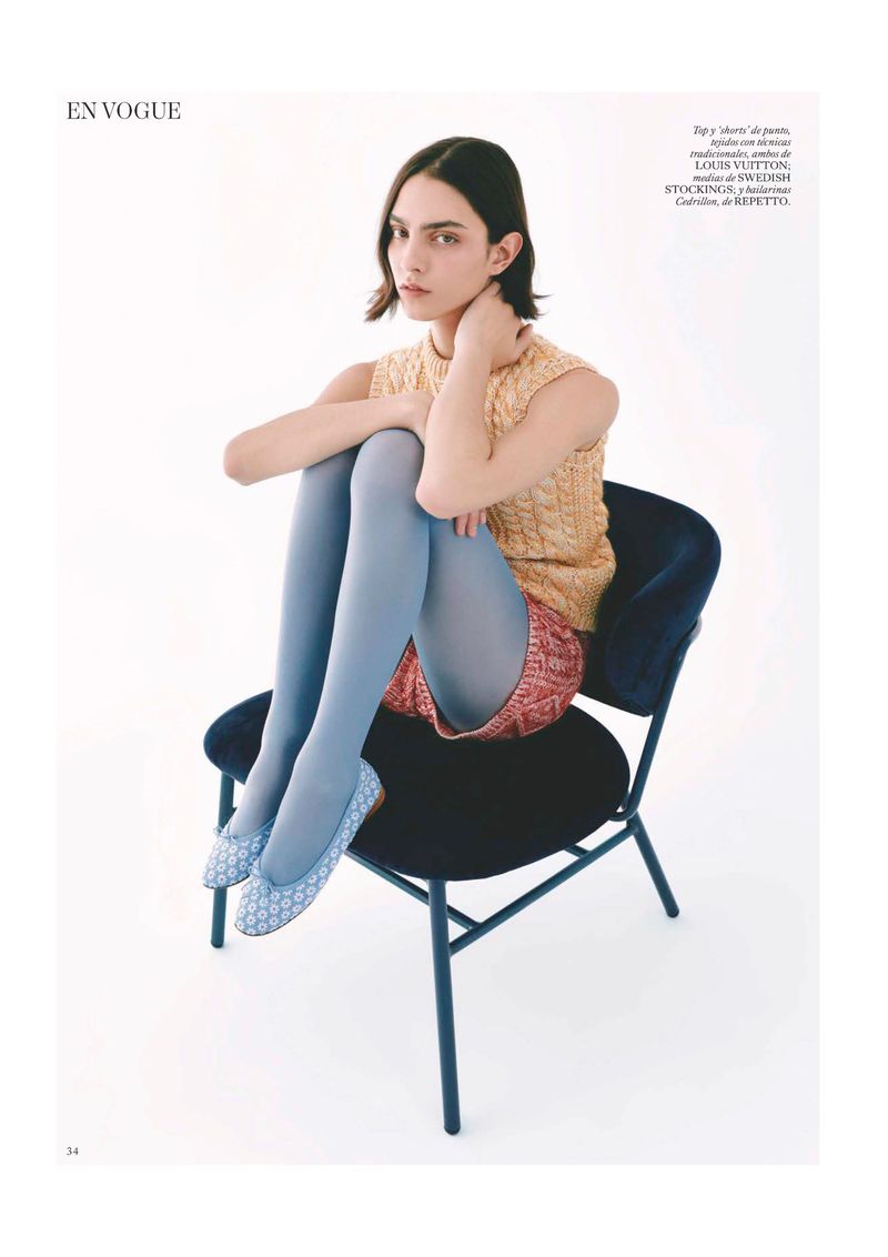 A young woman in a sleeveless sweater, short shorts, and blue tights sits on a blue chair against a white backdrop in a studio setting.