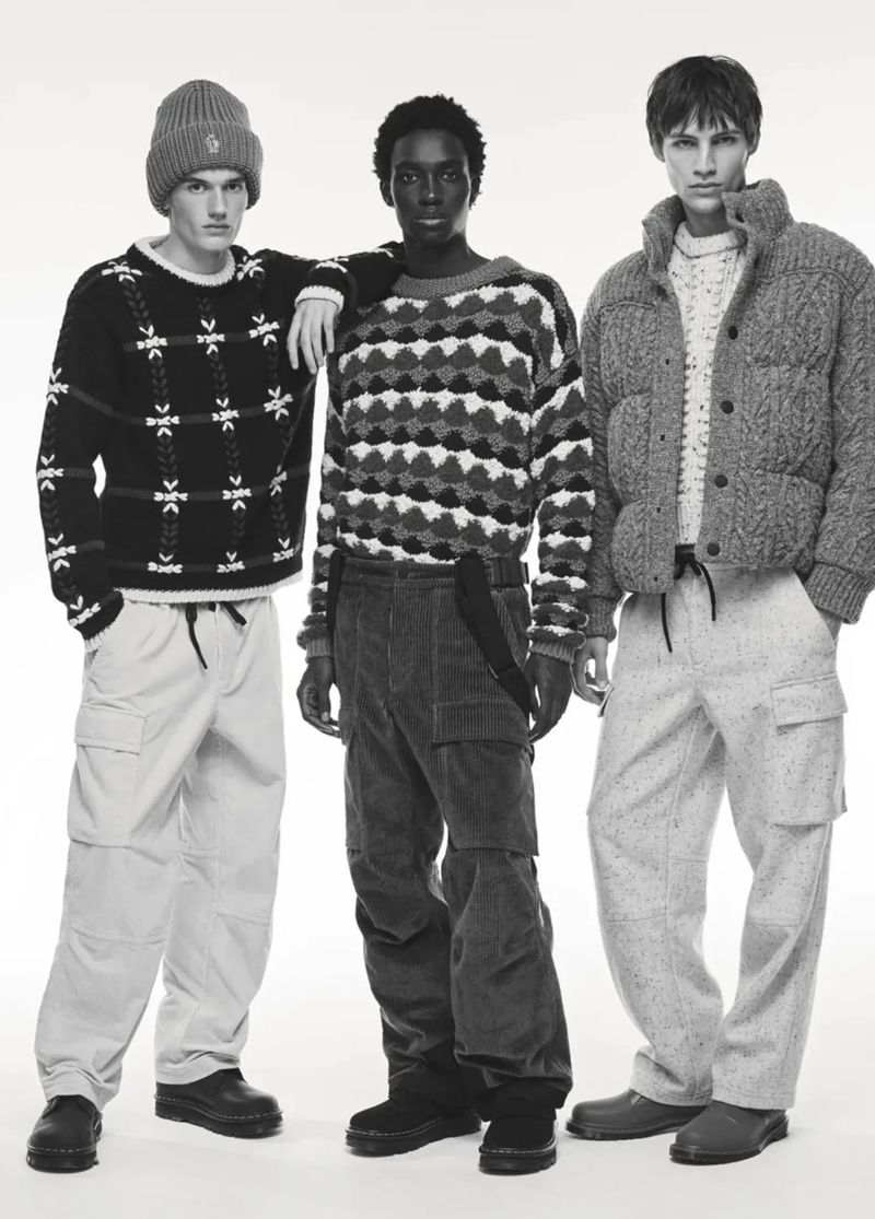 Three young adult men stand in a studio wearing patterned sweaters and loose trousers. One has a beanie, and the background is white.