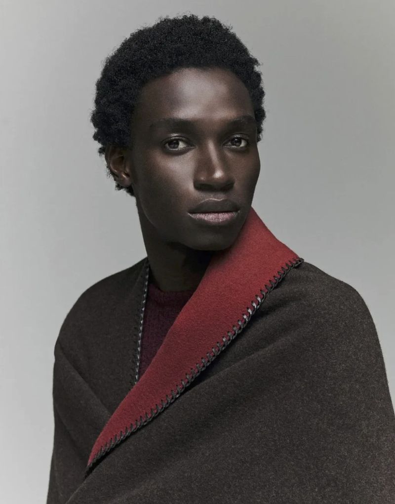 Portrait of a Black male fashion model wearing a brown and red shawl draped over his shoulders in a neutral studio setting.