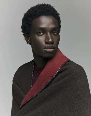 Portrait of a Black male fashion model wearing a brown and red shawl draped over his shoulders in a neutral studio setting.