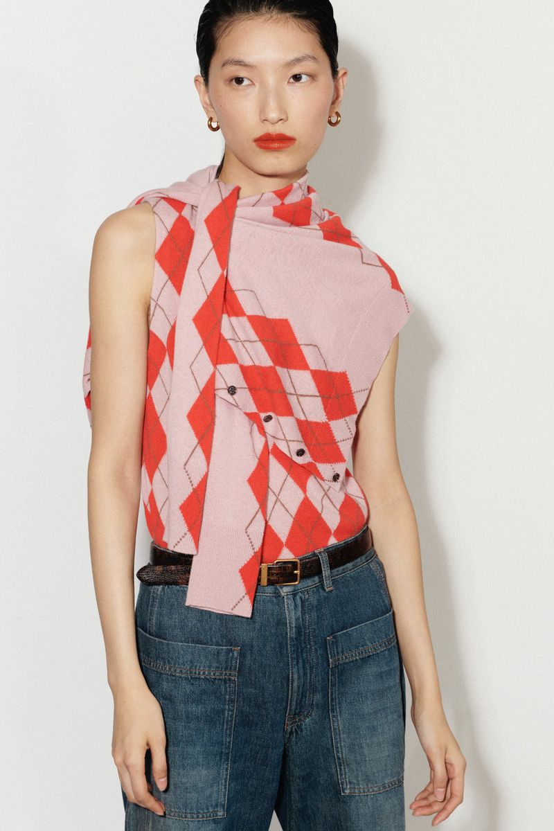 An Asian woman poses in blue jeans, a brown belt, and an argyle patterned sweater-scarf combination against a bright white background.