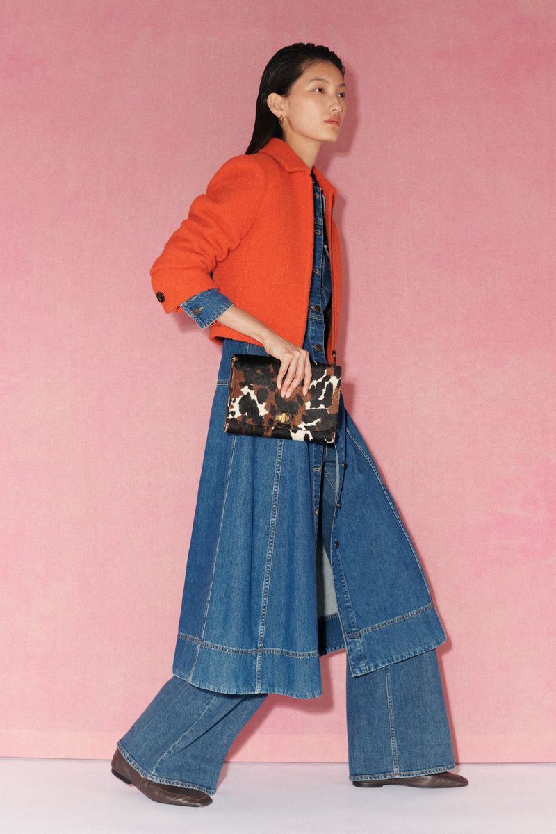 A stylish Asian woman wearing a denim dress and orange jacket, walking in a pink studio, holding a patterned clutch.