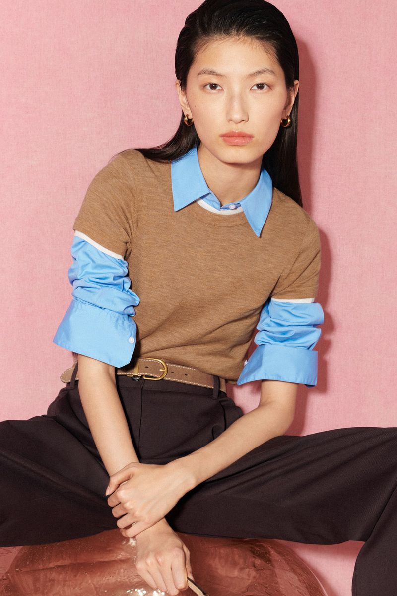 An Asian woman sits in a relaxed pose wearing a brown sweater, blue collared shirt, and black pants against a pink backdrop.