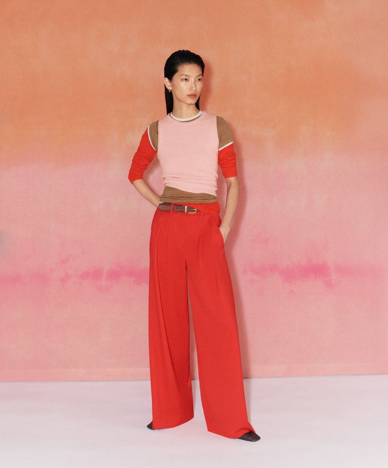 Asian fashion model poses in a pink top, red pants, and a brown belt against a gradient peach to pink background in a fashion editorial.