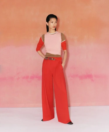 Asian fashion model poses in a pink top, red pants, and a brown belt against a gradient peach to pink background in a fashion editorial.