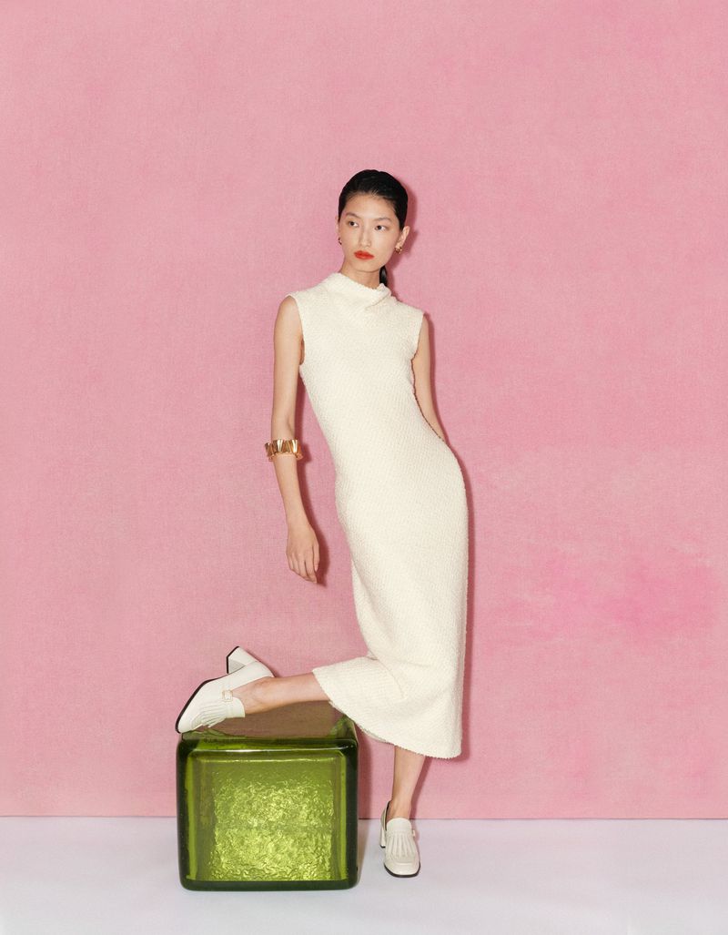 Stylish Asian woman poses in a white dress and loafers, leaning on an olive-green cube against a pink backdrop in a fashion shot.