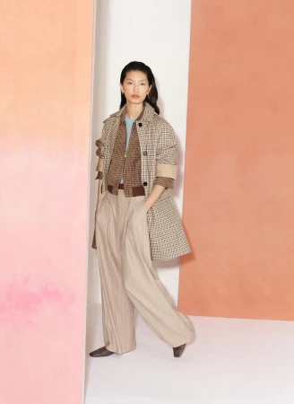 A young Asian woman poses in a beige patterned jacket, brown sweater, and tan pleated wide-leg trousers against a color-block backdrop.